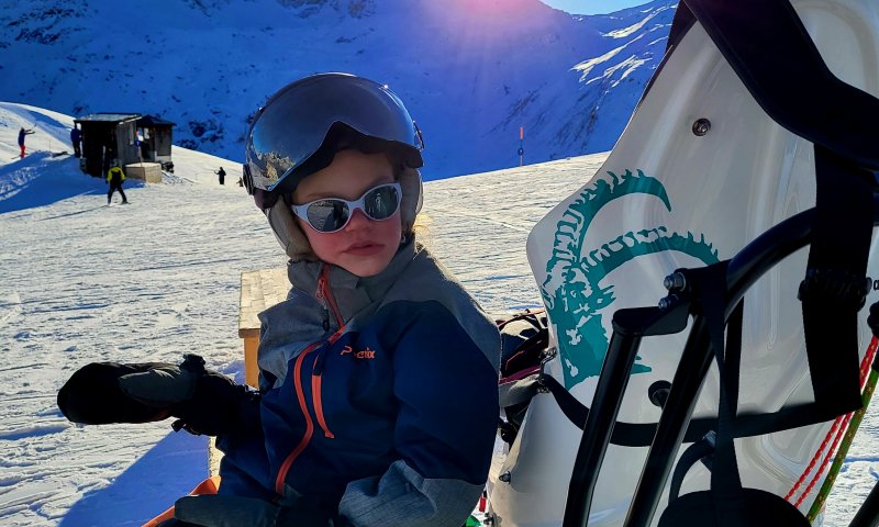 Ein Kind mit Beeinträchtigung sitzt auf einem Bänkli an der Sonne neben ihrem Dualski.