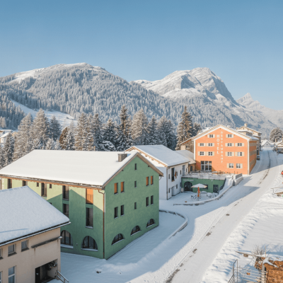 In einer Bergregion stehen bunte Häuser. Schnee bedeckt die Dächer und den Boden. Die Sonne scheint am blauen Himmel.