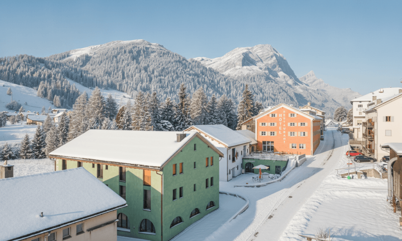 In einer Bergregion stehen bunte Häuser. Schnee bedeckt die Dächer und den Boden. Die Sonne scheint am blauen Himmel.