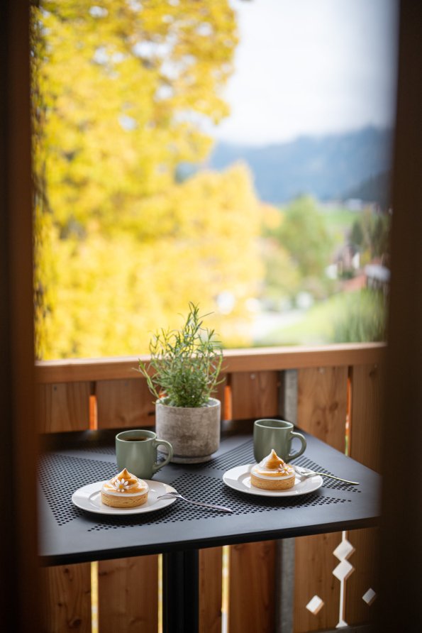 Auf einem Balkon stehen zwei Tassen Kaffee und zwei Kuchenstücke. Im Hintergrund leuchten die bunten Herbstblätter.
