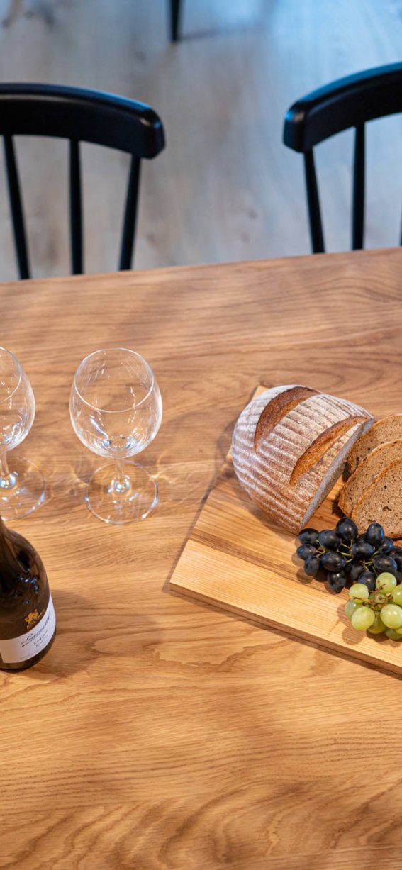 Auf einem Holztisch liegen Brot, Käse, Trauben und ein Weinflasche. Der Raum ist warm und einladend.