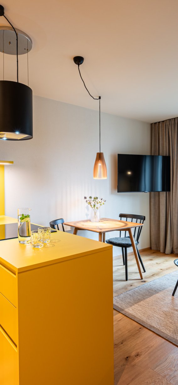 In einem hellen Raum stehen eine gelbe Küche und ein gemütlicher Wohnbereich mit Blick ins Freie.
