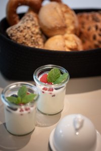 Zwei Gläser mit Joghurt, frischen Beeren und Minze stehen neben einer Auswahl an Brötchen auf einem Tisch.