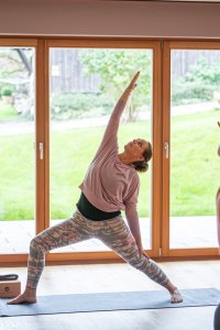 Drei Frauen praktizieren Yoga in einem hellen Raum mit großen Fenstern und Blick auf den Garten.