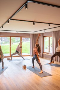 Fünf Personen üben Yoga in einem modernen Raum mit großen Fenstern. Die Atmosphäre ist ruhig und einladend.