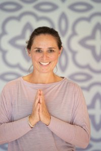 Eine Frau steht in einem hellen Raum mit einer entspannenden Pose und lächelt. Es ist eine ruhige Atmosphäre mit einem gemusterten Hintergrund.