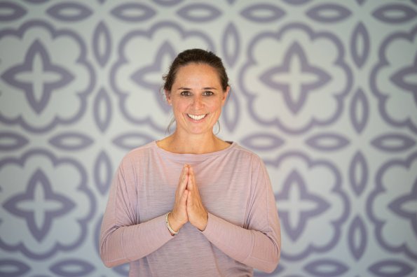 Eine Frau steht in einem hellen Raum mit einer entspannenden Pose und lächelt. Es ist eine ruhige Atmosphäre mit einem gemusterten Hintergrund.