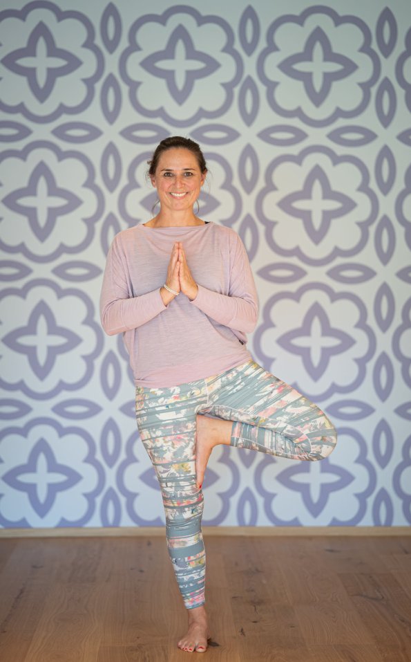 Eine Person macht einen Baum-Yoga-Stil in einem hellen Raum mit schöner Tapete. Sie lächelt und sucht nach Balance.