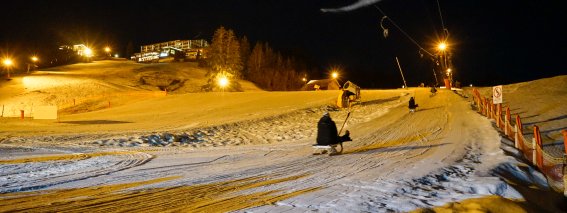 Mehrere Menschen rodeln mit Schlitten eine beleuchtete Piste hinunter. Es ist Nacht und der Schnee glitzert.