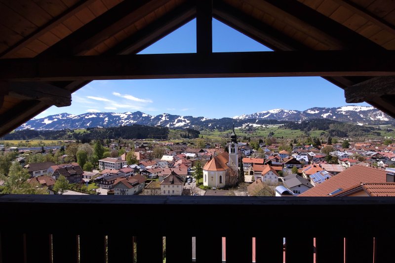Ausblick Balkon Himmeleck II Die Aussicht zeigt ein Dorf mit einem weißen Kirchengebäude. Umgeben von Bergen an einem sonnigen Tag. Es zeigt den Ausblick vom Balkon der Wohnung Himmeleck.