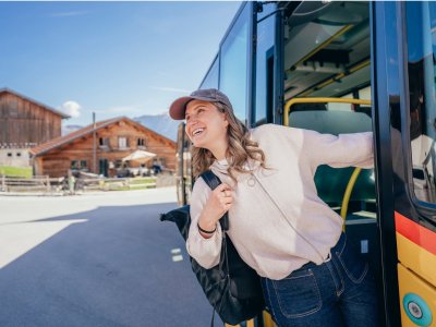 Eine Frau steigt aus einem Bus. Im Hintergrund sind Berge und ein Holzhaus zu sehen. Es ist ein sonniger Tag.
