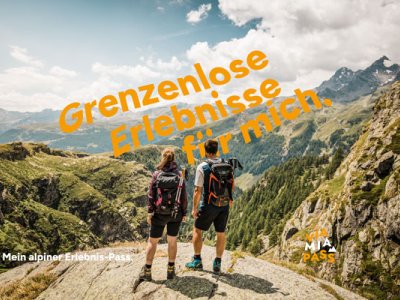 Zwei Personen stehen auf einem Gipfel und blicken auf die Berge. Es ist Tag und die Aussicht ist weit.