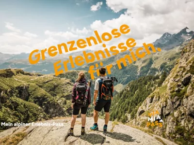 Zwei Personen stehen auf einem Gipfel und blicken auf die Berge. Es ist Tag und die Aussicht ist weit.