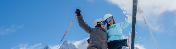 Zwei Personen fahren Ski in den Alpen. Sie lachen und genießen den Schnee an einem sonnigen Tag.
