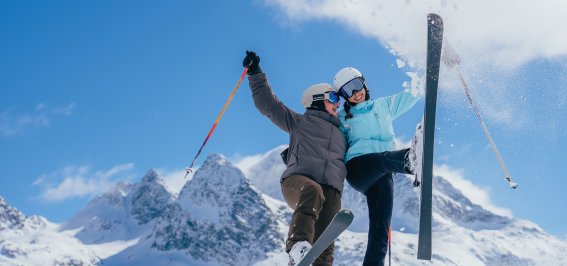 Zwei Personen fahren Ski in den Alpen. Sie lachen und genießen den Schnee an einem sonnigen Tag.