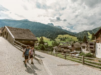 Zwei Wanderer gehen auf einem Weg in den Bergen. Die Sonne scheint und die Landschaft ist grün.