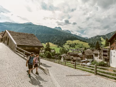 Zwei Wanderer gehen auf einem Weg in den Bergen. Die Sonne scheint und die Landschaft ist grün.