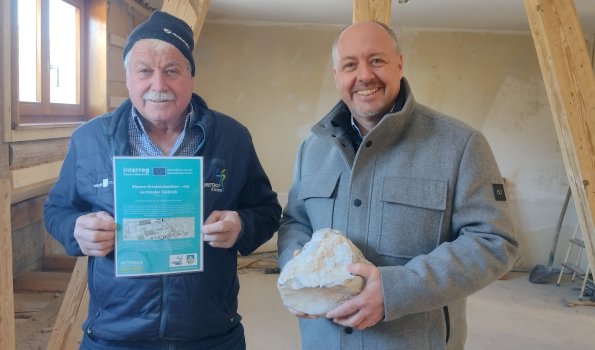 Zwei Männer stehen in einem leeren Raum. Sie halten ein Dokument und einen Stein in den Händen.