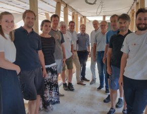 Eine Gruppe von Menschen steht für ein Foto in einem Halbkreis inmitten einer Baustelle. Im Hintergrund erkennt man Holzpflöcke.