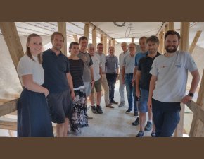 Eine Gruppe von Menschen steht für ein Foto in einem Halbkreis inmitten einer Baustelle. Im Hintergrund erkennt man Holzpflöcke.