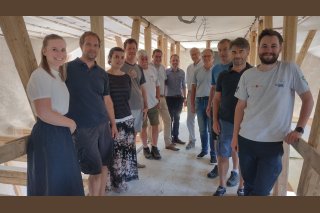 Info-Workshop Teilnehmer Eine Gruppe von Menschen steht für ein Foto in einem Halbkreis inmitten einer Baustelle. Im Hintergrund erkennt man Holzpflöcke.