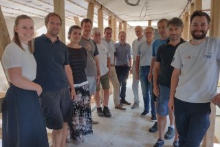 Info-Workshop Teilnehmer Eine Gruppe von Menschen steht für ein Foto in einem Halbkreis inmitten einer Baustelle. Im Hintergrund erkennt man Holzpflöcke.