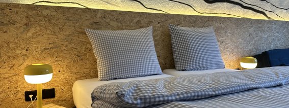 In einem Raum stehen zwei Betten mit karierten Bettbezügen. Lampen leuchten sanft und die Wände zeigen eine interessante Struktur.