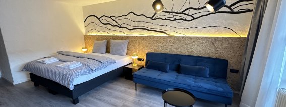 In einem Schlafzimmer gibt es ein Bett und ein Sofa. Die Wände sind schlicht dekoriert. Es gibt verschiedene Lampen.