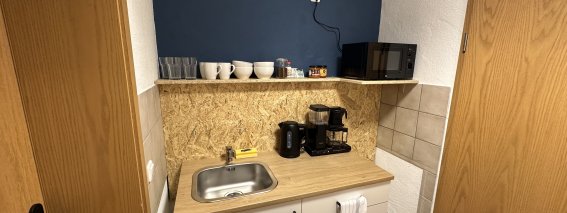 In einer kleinen Küche stehen Geräte, Geschirr und eine Holzarbeitsplatte an der Wand. Es ist tagsüber und hell.