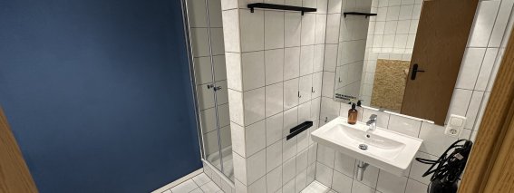 Ein Raum mit weißen Fliesen und einem Waschbecken. Es gibt eine Dusche und eine blaue Wand. Alles sieht neu aus.