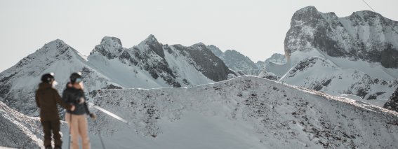 Zwei Skifahrer stehen auf der Piste und betrachten die beeindruckenden schneebedeckten Berge in den Alpen.
