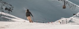 Ein Skifahrer fährt über den frischen Schnee in den Bergen. Die Gondel schwebt in der Ferne. Es ist ein klarer, sonniger Tag.
