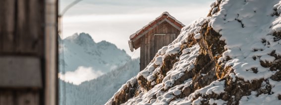 Ein altes Holzhaus steht in einer verschneiten Winterlandschaft. Die Berge im Hintergrund sind leicht bewölkt und bedeckt.