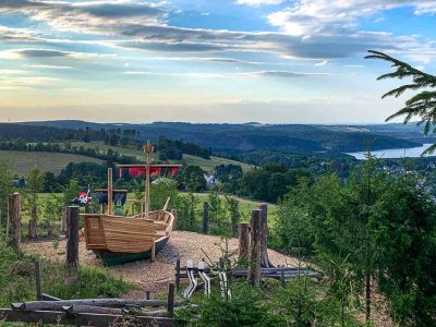 Ein Spielplatz mit einem Holzboot steht auf einer Wiese. Die Landschaft zeigt Hügel und einen See im Hintergrund.