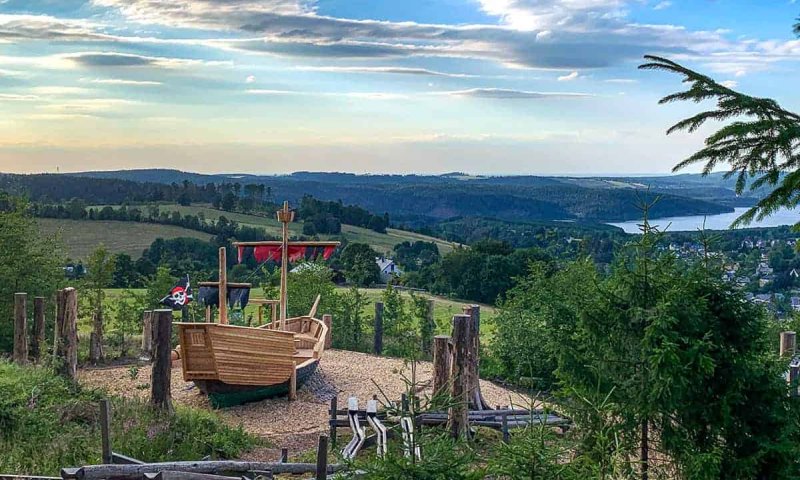 Ein Spielplatz mit einem Holzboot steht auf einer Wiese. Die Landschaft zeigt Hügel und einen See im Hintergrund.