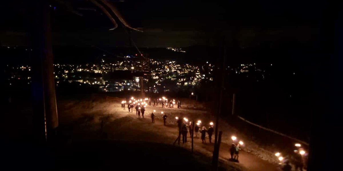 Fackelwanderung Eibenstock Eine Gruppe von Menschen wandert mit Fackeln durch die dunkle Berglandschaft, beleuchtet von funkelnden Lichtern in der Ferne.