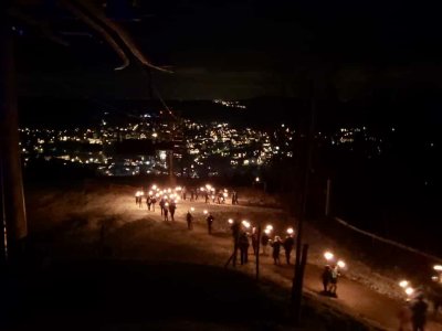 Eine Gruppe von Menschen wandert mit Fackeln durch die dunkle Berglandschaft, beleuchtet von funkelnden Lichtern in der Ferne.