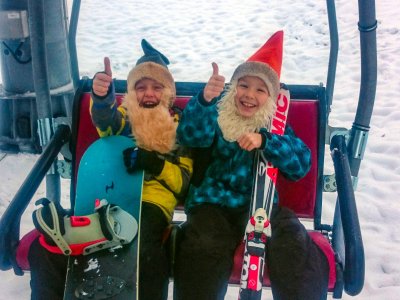 Zwei Kinder sitzen im Skilift mit Snowboard und Skiern. Beide zeigen begeistert ihren Daumen nach oben.