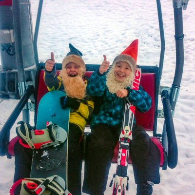 Zwei Kinder sitzen im Skilift mit Snowboard und Skiern. Beide zeigen begeistert ihren Daumen nach oben.