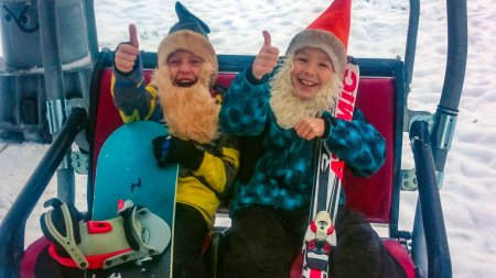 Zwei Kinder sitzen im Skilift mit Snowboard und Skiern. Beide zeigen begeistert ihren Daumen nach oben.