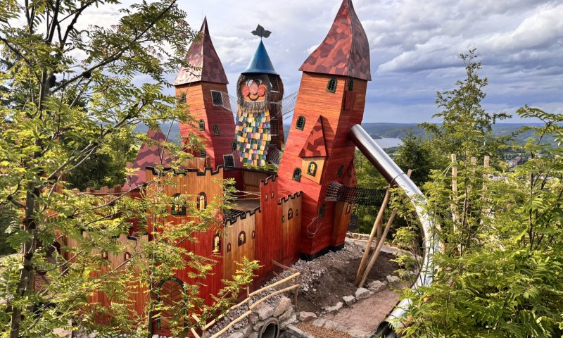 Kinder spielen fröhlich in einer bunten Burg mit Rutsche, umgeben von Bäumen und grauem Himmel.