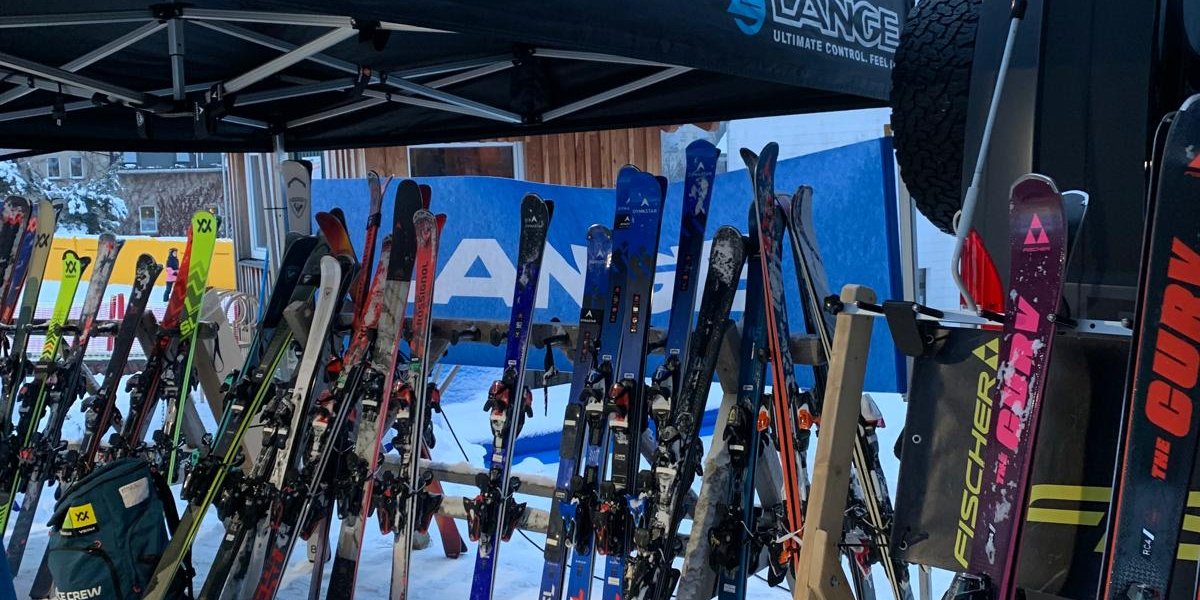 Auf einem Wintermarkt stehen verschiedene Skier unter einem Zelt. Es ist taghell und die Atmosphäre ist lebendig und einladend.