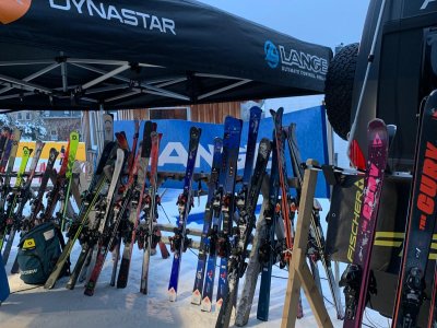 Auf einem Wintermarkt stehen verschiedene Skier unter einem Zelt. Es ist taghell und die Atmosphäre ist lebendig und einladend.