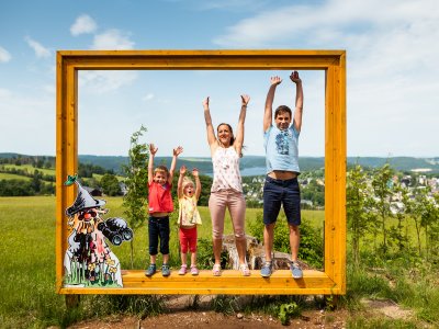 Eine Familie posiert fröhlich vor einem großen Bilderrahmen in der Natur. Es ist ein schöner Sonnentag bei Wurzelrudi im Sommer.