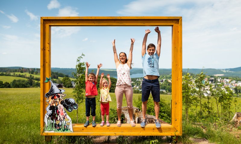 Eine Familie posiert fröhlich vor einem großen Bilderrahmen in der Natur. Es ist ein schöner Sonnentag bei Wurzelrudi im Sommer.