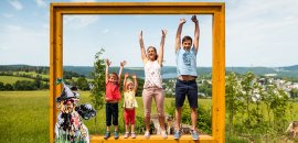 Eine Familie mit zwei Kindern posiert im Freien vor einem großen Holzrahmen mit einem bunten Motiv und genießt den Tag.