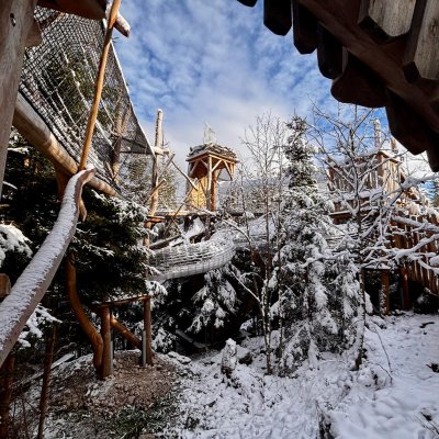 Schnee bedeckt die Bäume und Strukturen eines Waldspielplatzes an einem klaren Wintertag. Kinder können spielen und erkunden.
