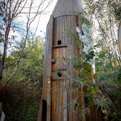 Ein Holz-Spielturm steht im Freien umgeben von Bäumen und Pflanzen. Kinder können darin spielen und erkunden.