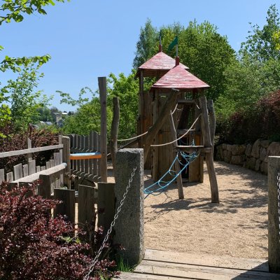 Kinder spielen auf einem Spielplatz mit Holzgeräten und Sand. Der Platz ist umgeben von Bäumen und strahlend blauem Himmel.