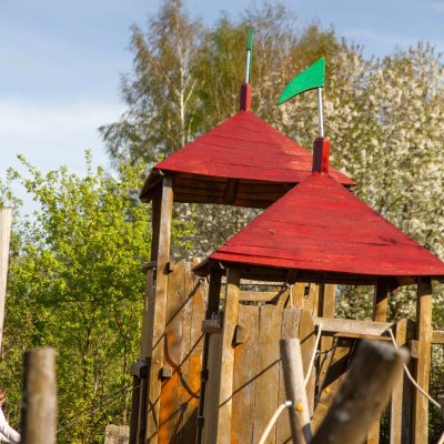 Kinder spielen auf einem Spielplatz mit Holzstrukturen. Die Sonne scheint und Bäume sind im Hintergrund zu sehen.